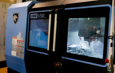 Maszyna CNC typu DNM 5700 pracuje z systemem rozpylania płynu chłodzącego wewnątrz obudowy, co można obserwować przez szklane okienko. Na zewnętrznej powierzchni maszyny umieszczono etykiety bezpieczeństwa i instrukcje obsługi.